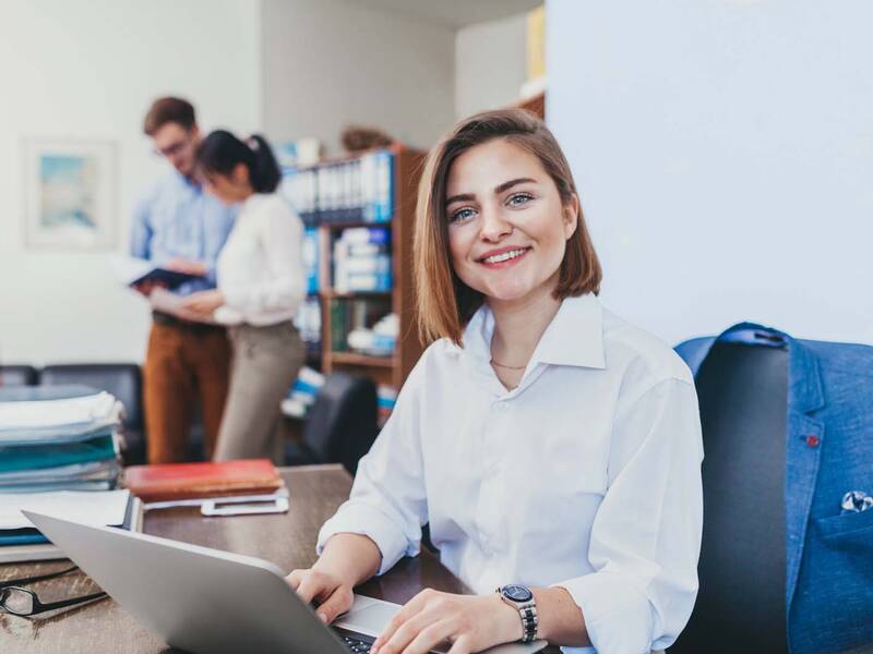 Eine Frau arbeitet an ihrem Laptop. Im Hintergrund ein Büro mit weiteren Personen.